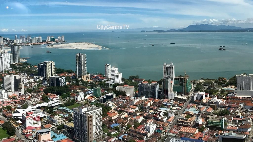 Penang Aerial view of a coastal city with a mix of high-rise buildings and traditional houses, overlooking the sea and distant mountains.